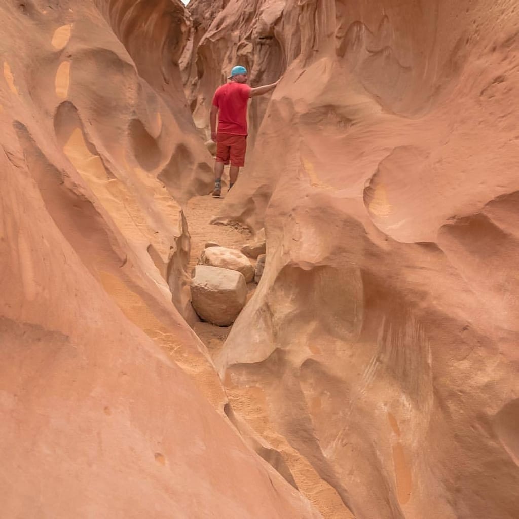 A hiker in a red shirt stands in a pink orange backcountry slot canyon in Southern Utah.