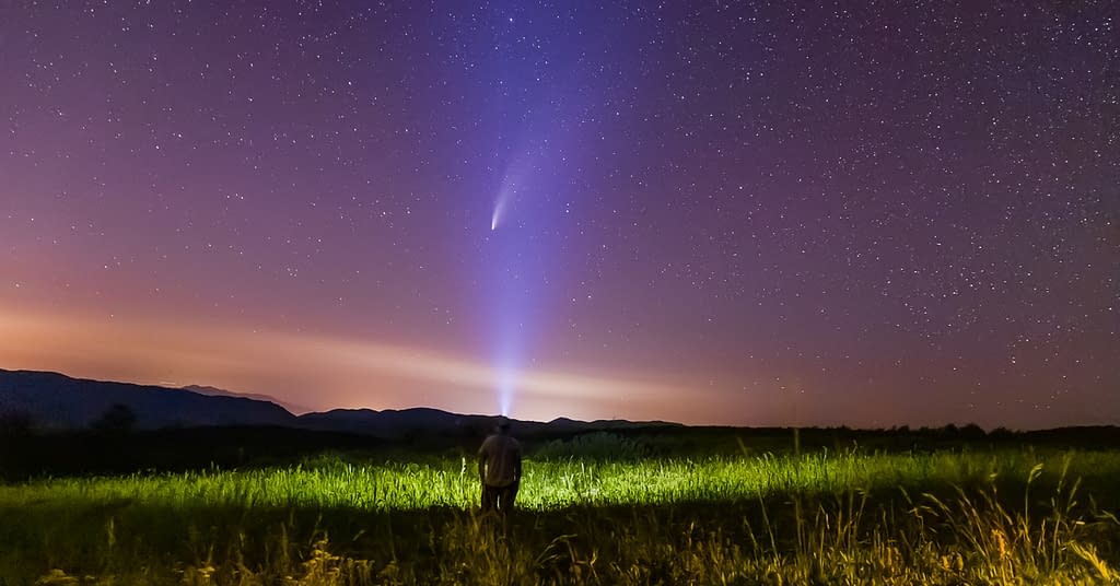 A man stands in a flower-filled meadow with a light shining towards a starry night sky as Comet Neowise passes over the mountain range in Northern Utah