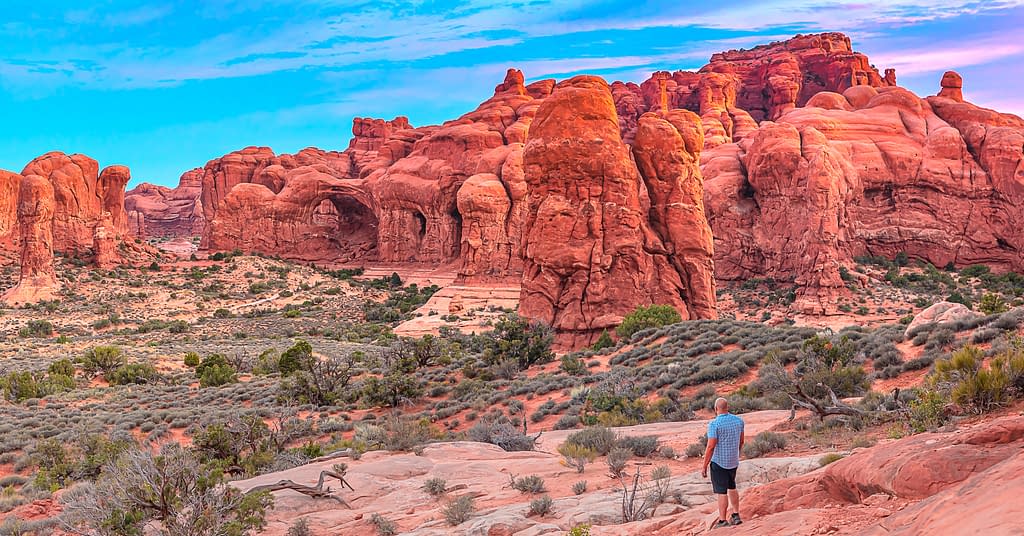 An outdoor explorer dressed in summer shorts and a blue shirt stands on a cliff admiring unique towering desert rock formations at sunrise in Arches National Park in Moab, Utah