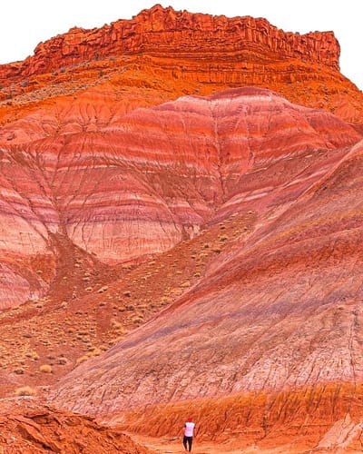 A man in a red hat stands at the bottom of a large colorful rainbow-striped mountain at Paria Canyon-Vermilion Cliffs Wilderness Area in Utah