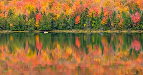 An outdoor explorer paddles a canoe among colorful autumn foliage as the vibrant pink colors reflect in Heart Lake in the Adirondack Region of Upstate New York