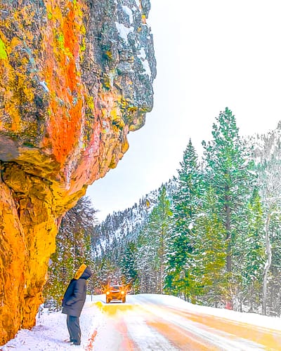 A man stands in the snow covered road in a forest looking up at a large and colorful mountain with lichens growing on it as an off-road truck approaches in the wintertime in Painted Rocks State Park in the West Fork Valley of the Bitterroot Mountains Near Hamilton, Montana
