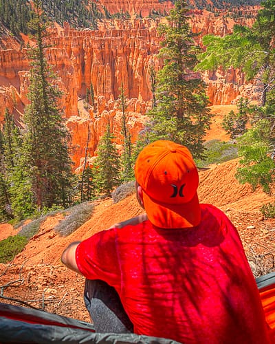 A man dressed in an orange hat and red shirt sits on a mountain in a hammock looking at scenic views of orange hoodoo rock formations, pink cliffs, and a forest of pine trees at Bryce Canyon National Park in Utah