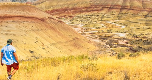 A man wearing colorful clothing walks on a hiking trail towards colorful desert hills in John Day Fossil Beds National Monument in Kimberly, Oregon