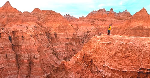 A man dressed in an orange jacket and striped winter hat stands at a scenic viewpoint on a hiking trail looking at colorful buttes, rock formations, and canyons at Badlands National Park in South Dakota