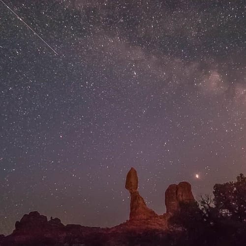A shooting star falls across the sky and The Milky Way at Balanced Rock in Arches National Park in Moab, Utah.