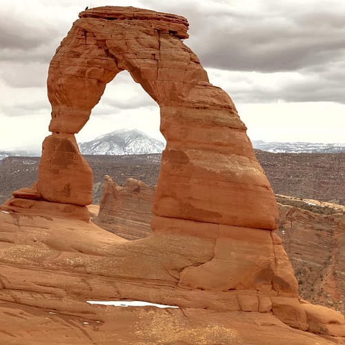 A snow-covered mountain is frame by an opening in a natural sandstone rock arch at Delicate Arch in Arches National Park