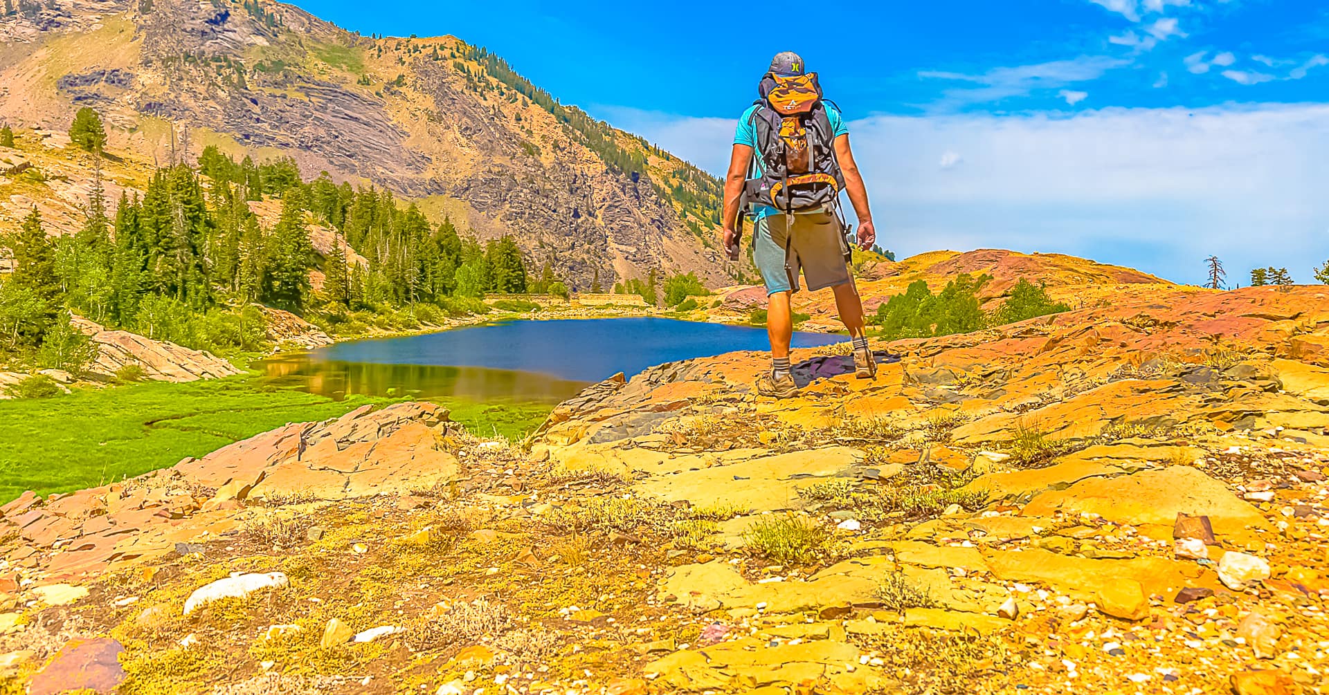 An outdoor backpacker wearing hiking gear stands on a mountain overlook looking at a beautiful blue lake surrounded by a pine tree forest at Lake Blanche in Big Cottonwood Canyon at the Twin Peaks Wilderness Area in Salt Lake City, Utah
