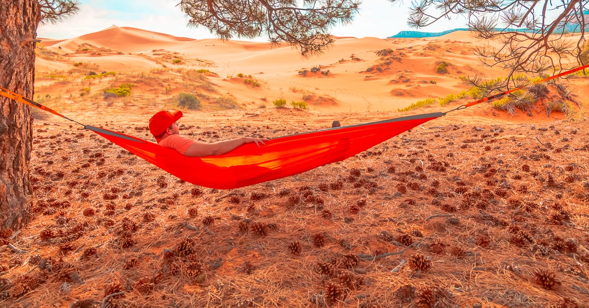 A man with an orange hat rests on an orange hammock under a tree looking at bright yellow desert sand dunes at Coral Pink Sand Dunes State Park in Kanab, Utah