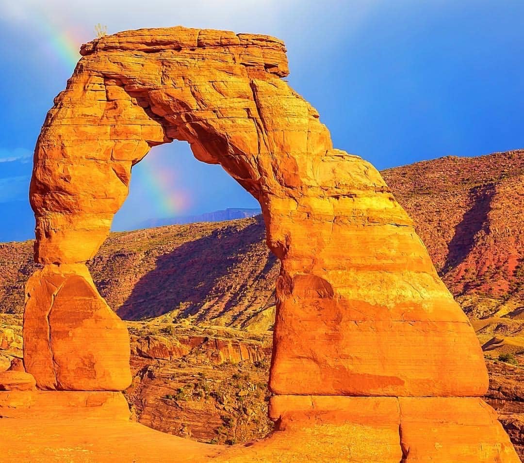 A colorful rainbow passes through Delicate Arch on a trail during monsoon season at Arches National Park in Moab, Utah.