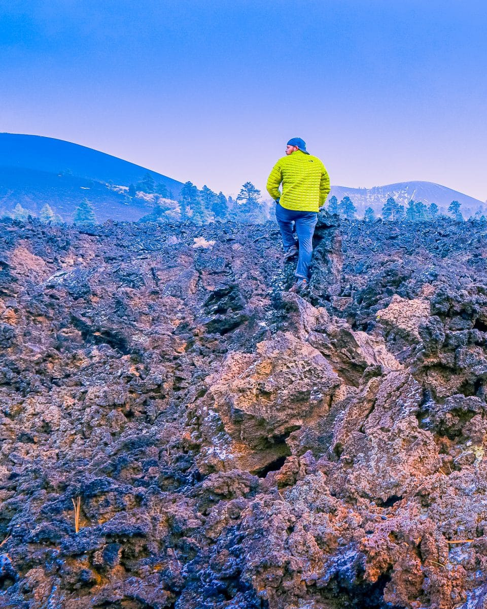 An outdoor explorer climbs colorful textured volcanic rock to view snowy mountain peaks and green forest pine trees at Sunset Crater Volcano National Monument Cinder Cone in Northern Arizona near Flagstaff, Arizona