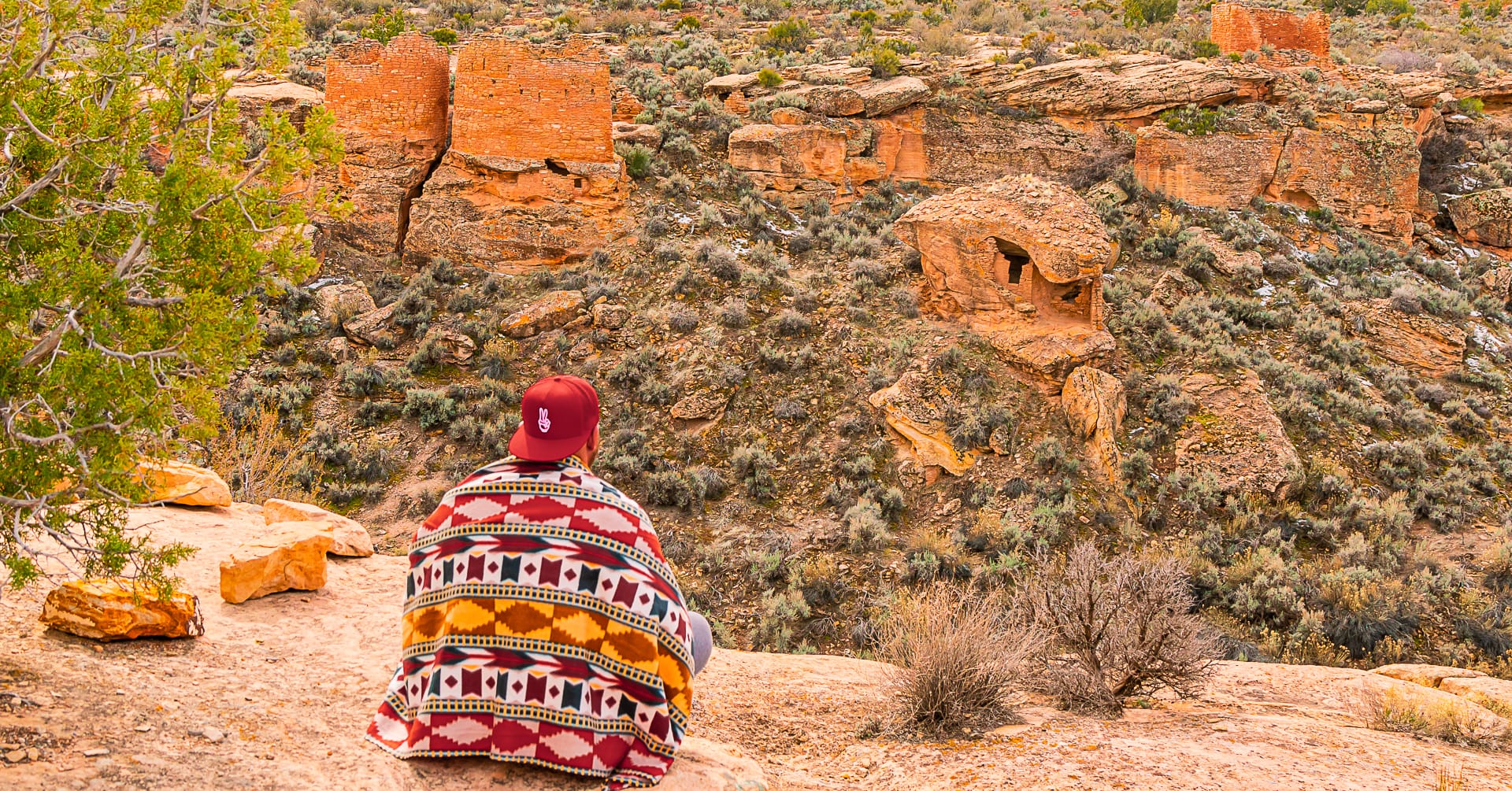 A man sits on a cliff wearing a red hat and wrapped in a colorful native-inspired blanket looking at ancient rock dwellings at Hovenweep National Monument in Blanding, Utah