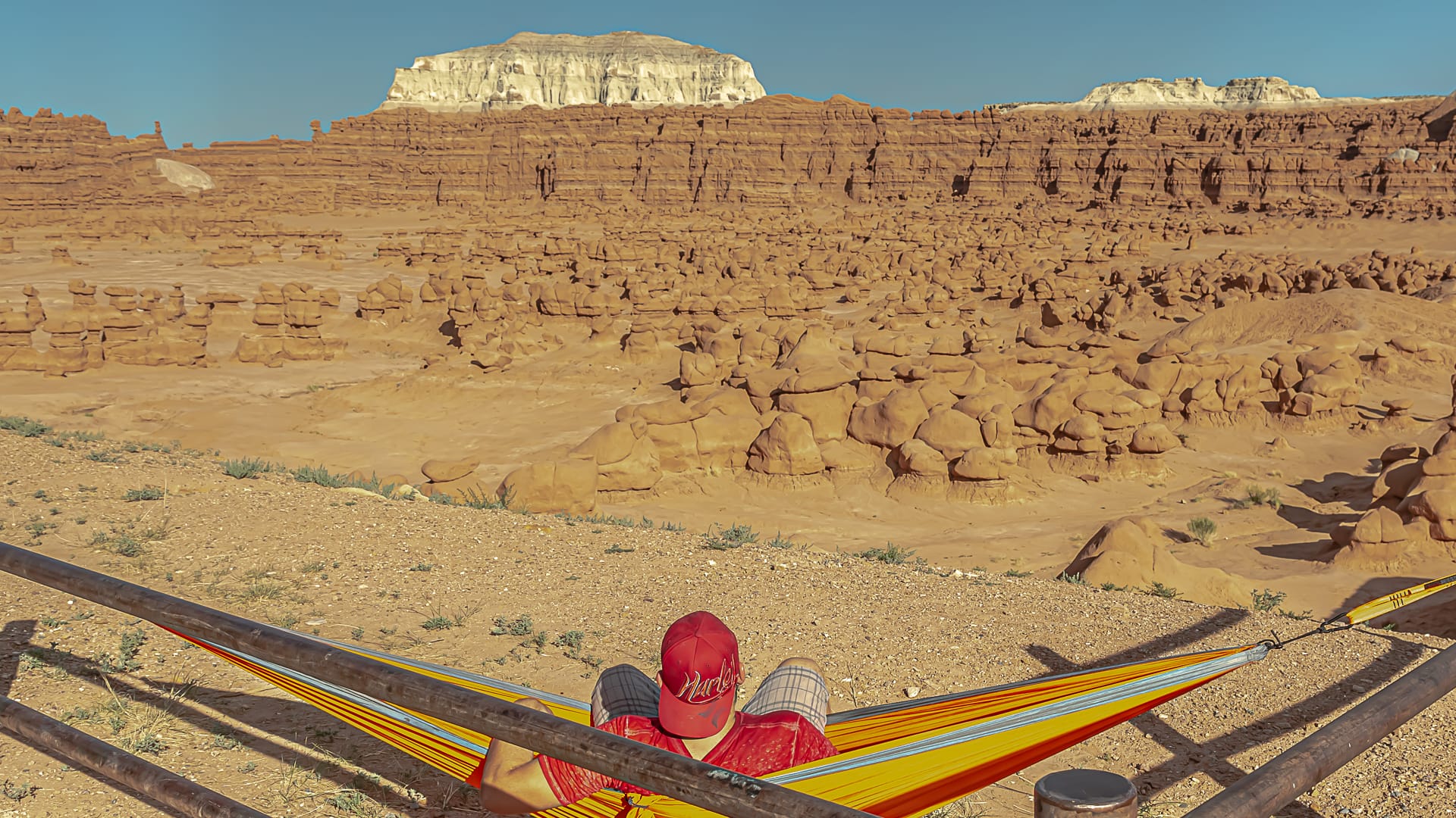 A man with a red hat and shirt sits in an outdoor hammock looking at unique rock formations and hoodoos at Goblin Valley State Park in Green River, Utah